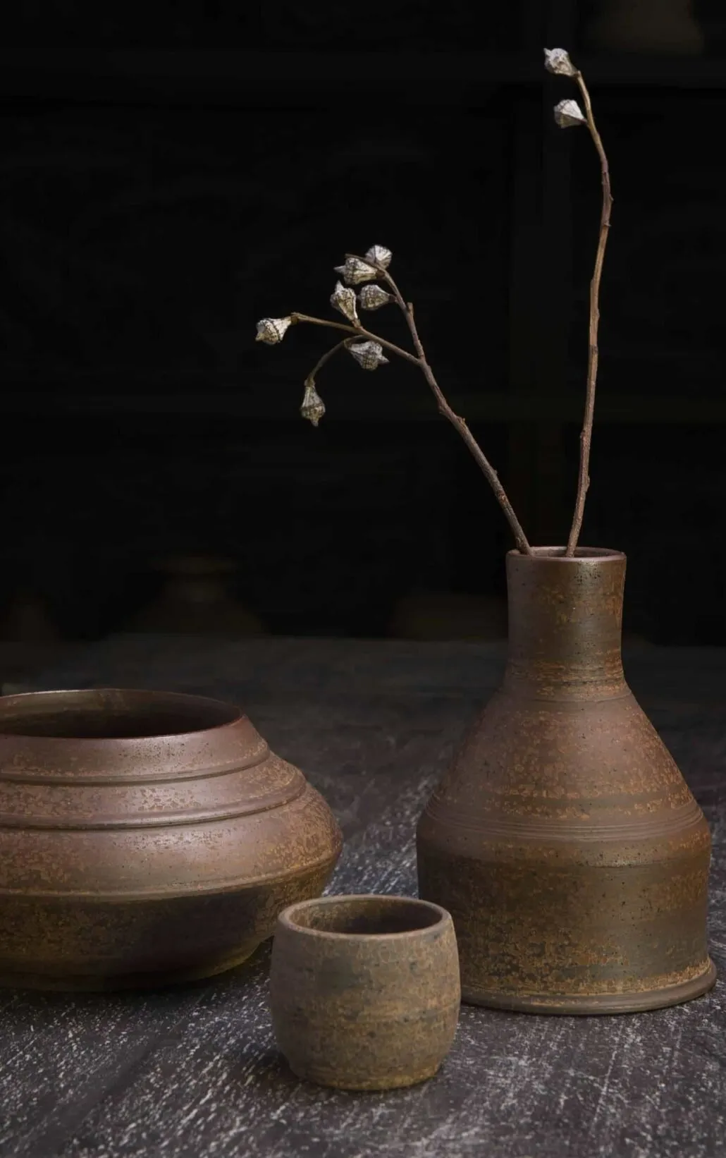 A slender vase with a single dried stem beside two small bowls